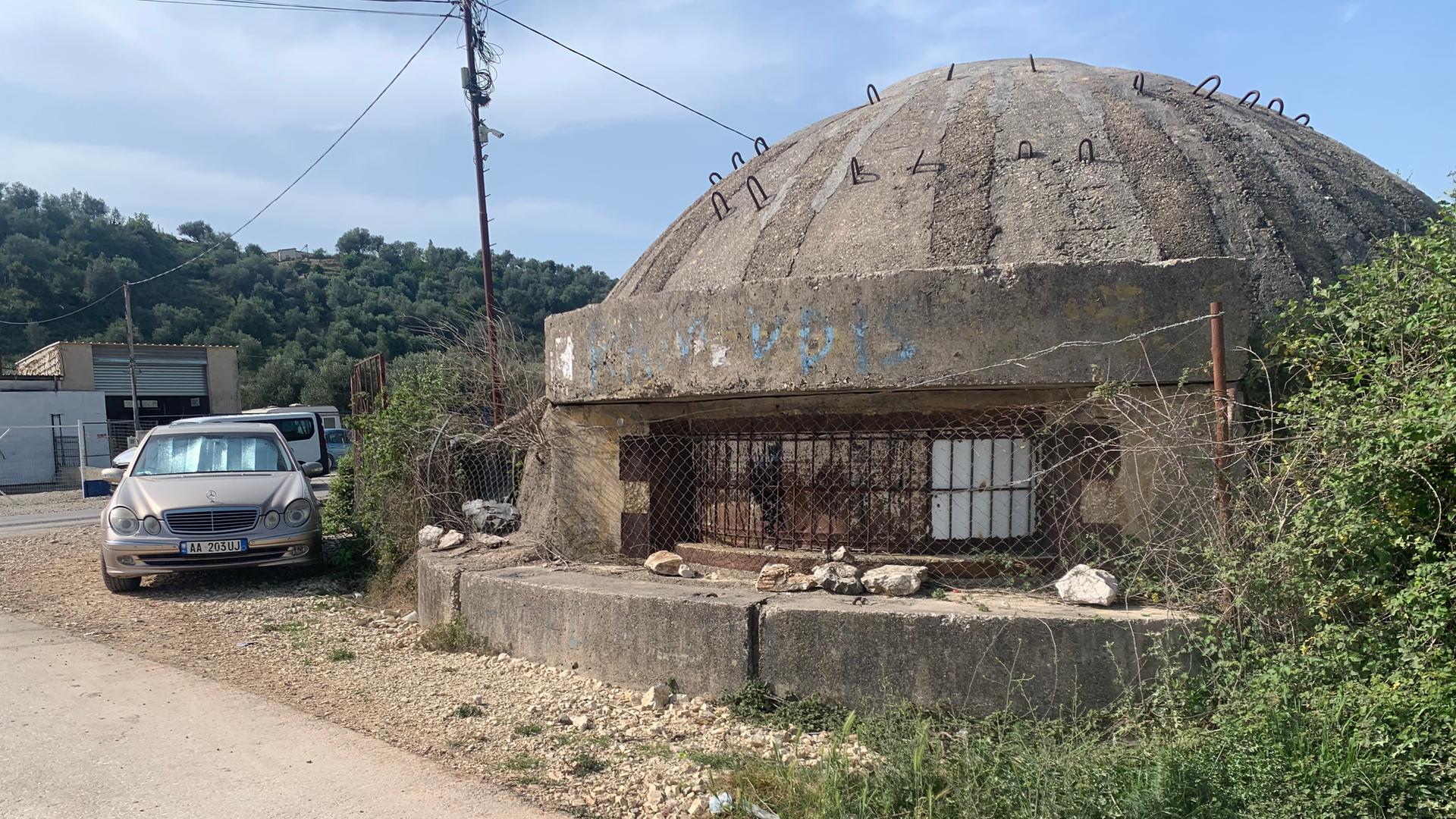 An einem Schotterweg steht ein großer Bunker aus Beton mit einem runden Dach. Der Bunker ist umgeben von Gestrüpp.