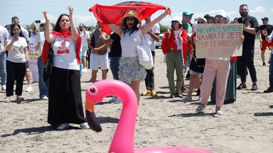 Albanien: Demonstrierende am Strand mit albanischer Flagge, Protestplakaten und Flamingo-Rettungsring.