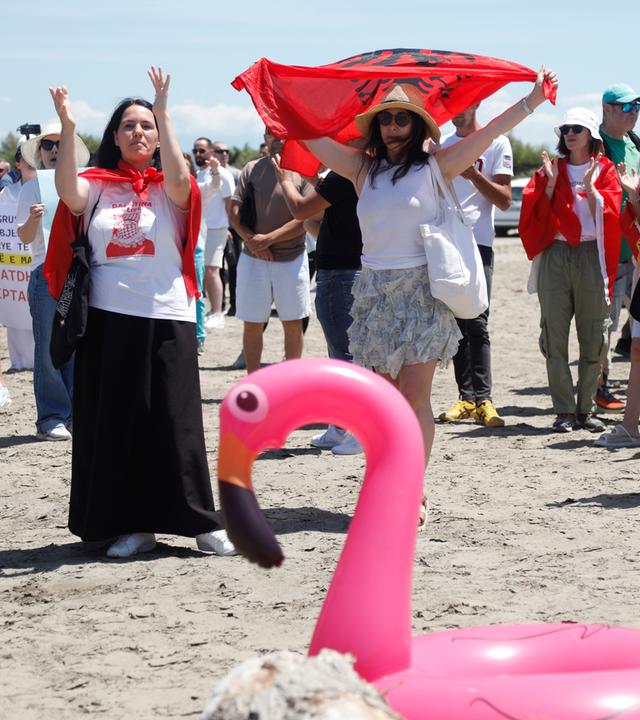 Albanien: Demonstrierende am Strand mit albanischer Flagge, Protestplakaten und Flamingo-Rettungsring.