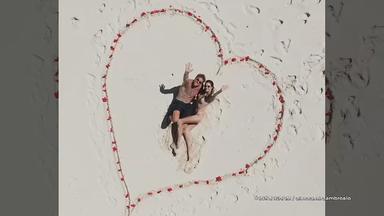 Alessandra Ambrosio und Buck Palmer liegen am Strand innerhalb eines mit roten Blüten gelegten Herzsymbols im Sand und blicken nach oben in die Kamera.