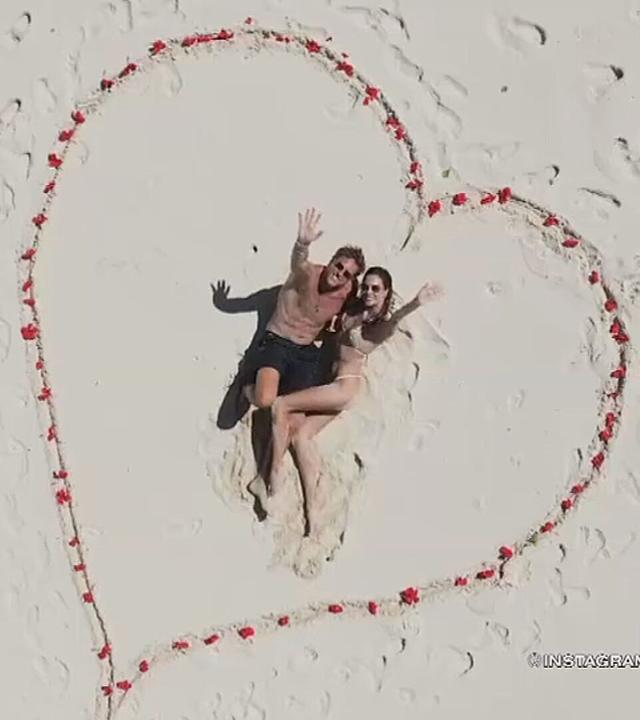 Alessandra Ambrosio und Buck Palmer liegen am Strand innerhalb eines mit roten Blüten gelegten Herzsymbols im Sand und blicken nach oben in die Kamera.