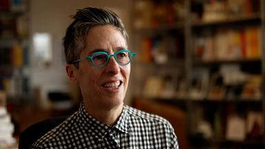 Porträtfoto von Alison Bechdel, einer Frau mit kurzen zurückgekämmten Haaren, die eine auffällige runde türkisfarbene Brille und ein schwarz-weiß-kariertes Hemd trägt.