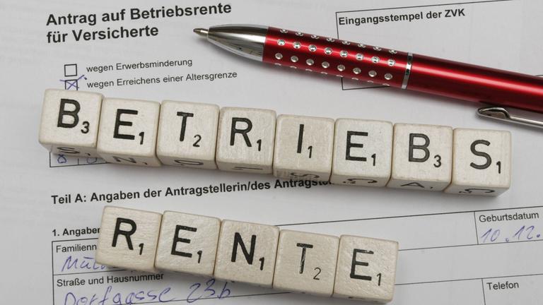 Auf einem Antragsformular für Betriebsrente liegen ein roter Kugelschreiber und Steine mit Buchstaben, die das Wort "Betriebsrente" bilden.