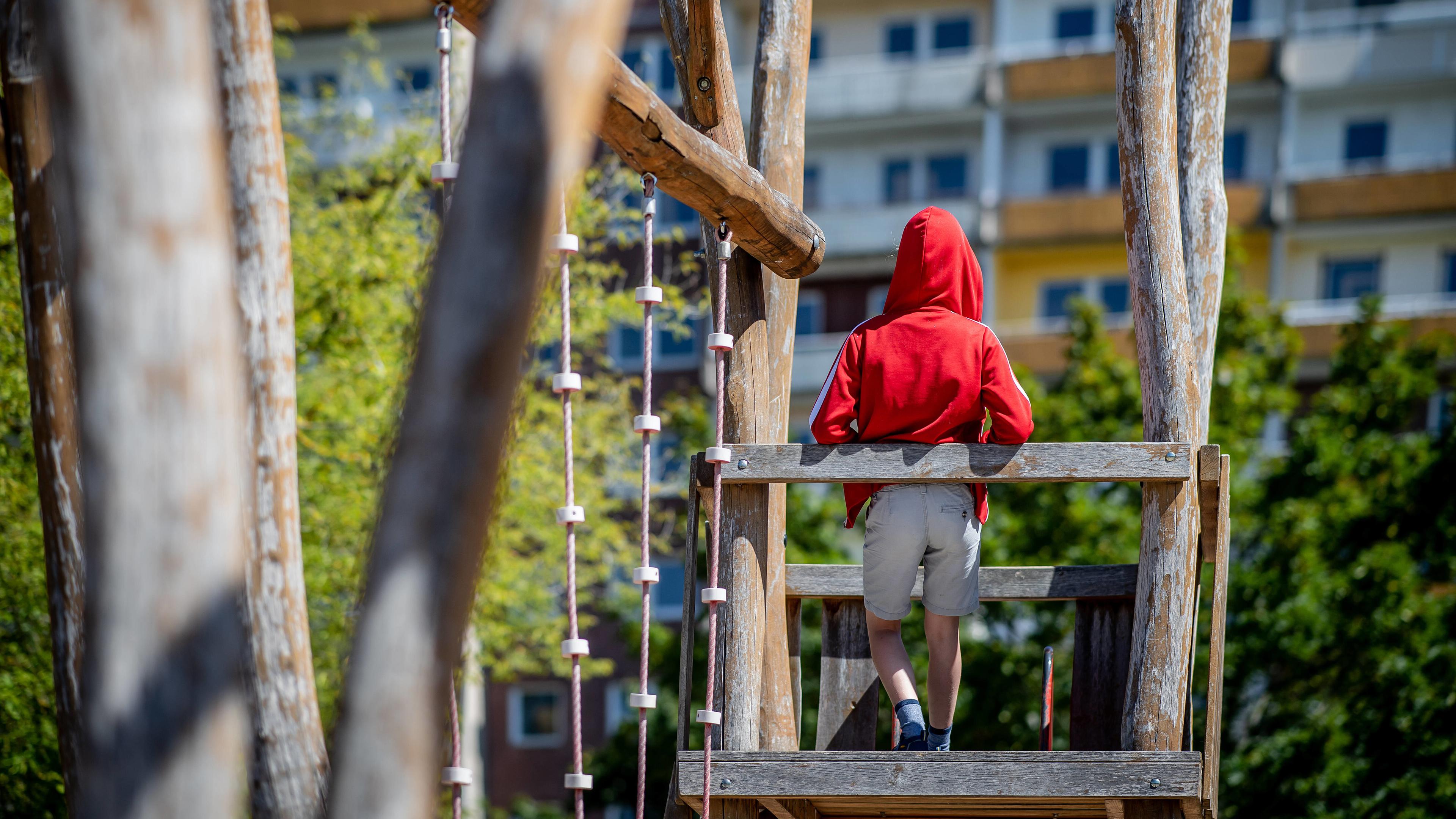 "ZDF.reportage: Alltag Kinderarmut - Kein Geld - keine Chance?": Ein Junge mit dem Rücken zur Kamera, mittig im Bild, auf einem Klettergerüst. Er trägt eine rote Kapuze und helle Shorts. Im Hintergrund sieht man einen Plattenbau.