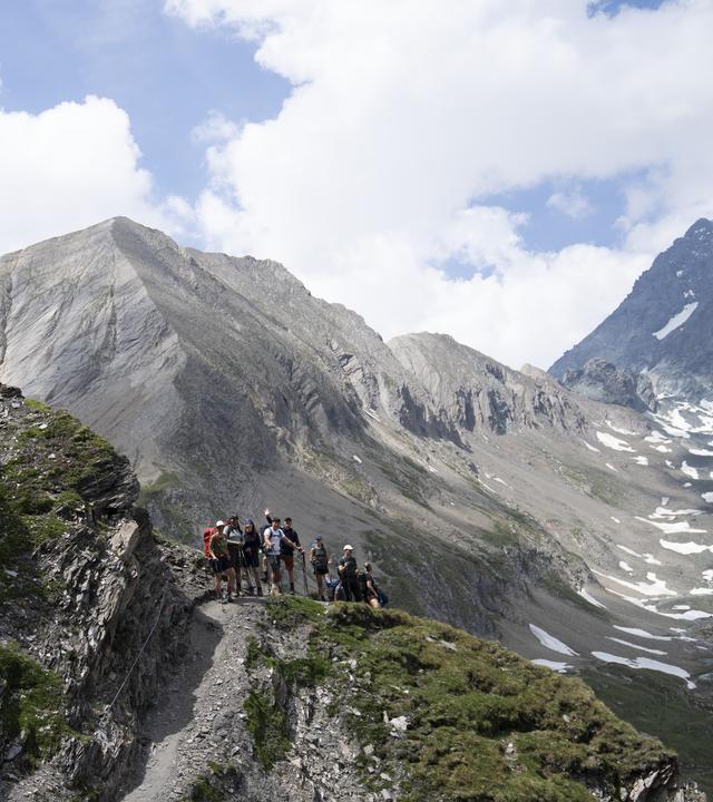 "Abenteuer Alpenüberquerung - Absturzgefahr: Lisa kämpft mit der Angst": Eine Wandergruppe folgt einem schmalen Pfad durch eine felsige Hochgebirgslandschaft. Der Weg führt an einem steilen, stellenweise begrünten Hang entlang. Im Hintergrund erheben sich graue Berggipfel mit Schneefeldern unter einem bewölkten Himmel.