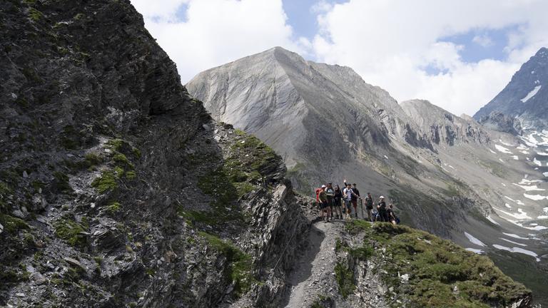"Abenteuer Alpenüberquerung - Absturzgefahr: Lisa kämpft mit der Angst": Eine Wandergruppe folgt einem schmalen Pfad durch eine felsige Hochgebirgslandschaft. Der Weg führt an einem steilen, stellenweise begrünten Hang entlang. Im Hintergrund erheben sich graue Berggipfel mit Schneefeldern unter einem bewölkten Himmel.