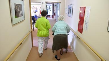 Eine Altenpflegerin begleitet eine Seniorin an einem Rollator durch einen Flur.