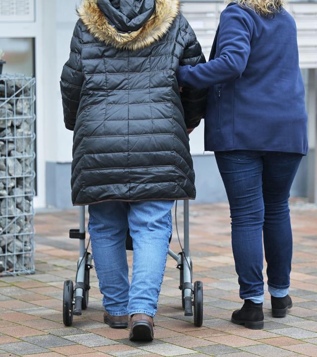 Eine Seniorin mit Rollator wird von einer Pflegedienstmitarbeiterin auf dem Weg zu ihrer Wohnung gestützt.