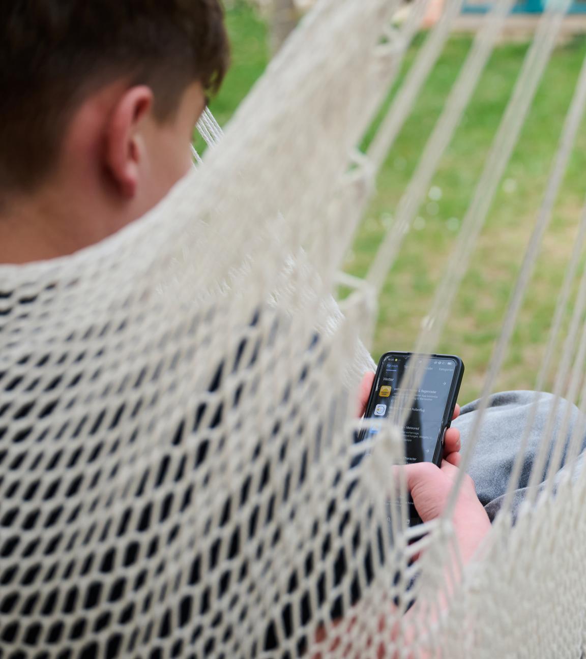  Ein Junge guckt auf sein Smartphone, während er in einer Hängeschaukel in einem Garten sitzt.