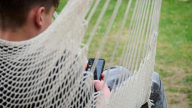  Ein Junge guckt auf sein Smartphone, während er in einer Hängeschaukel in einem Garten sitzt.
