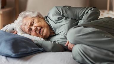 Eine ältere Frau liegt seitlich im Bett und versucht zu schlafen. Ihr Gesichtsaudruck deutet auf Schmerzen hin. 