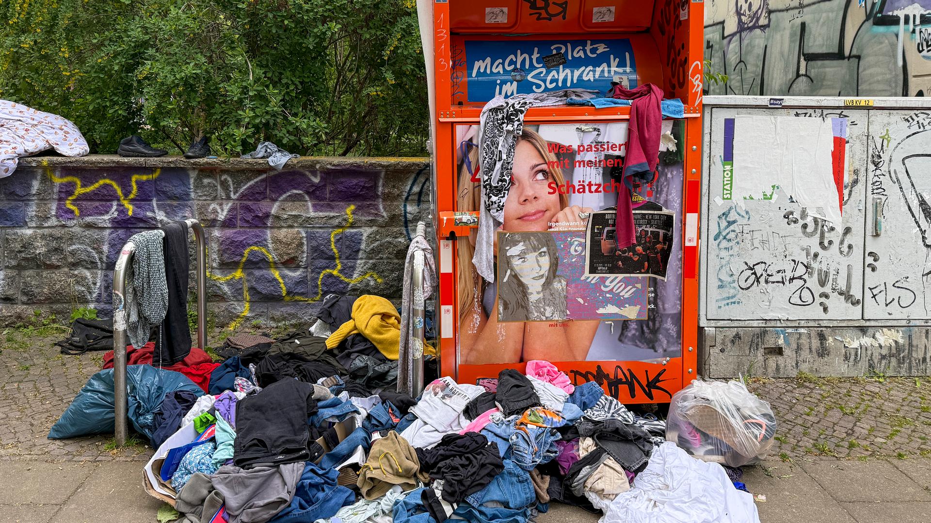 Symbolbild: Aufgerissene Müllsäcke und wild umhergeworfene Kleidung liegen vor einem Altkleidercontainer. 