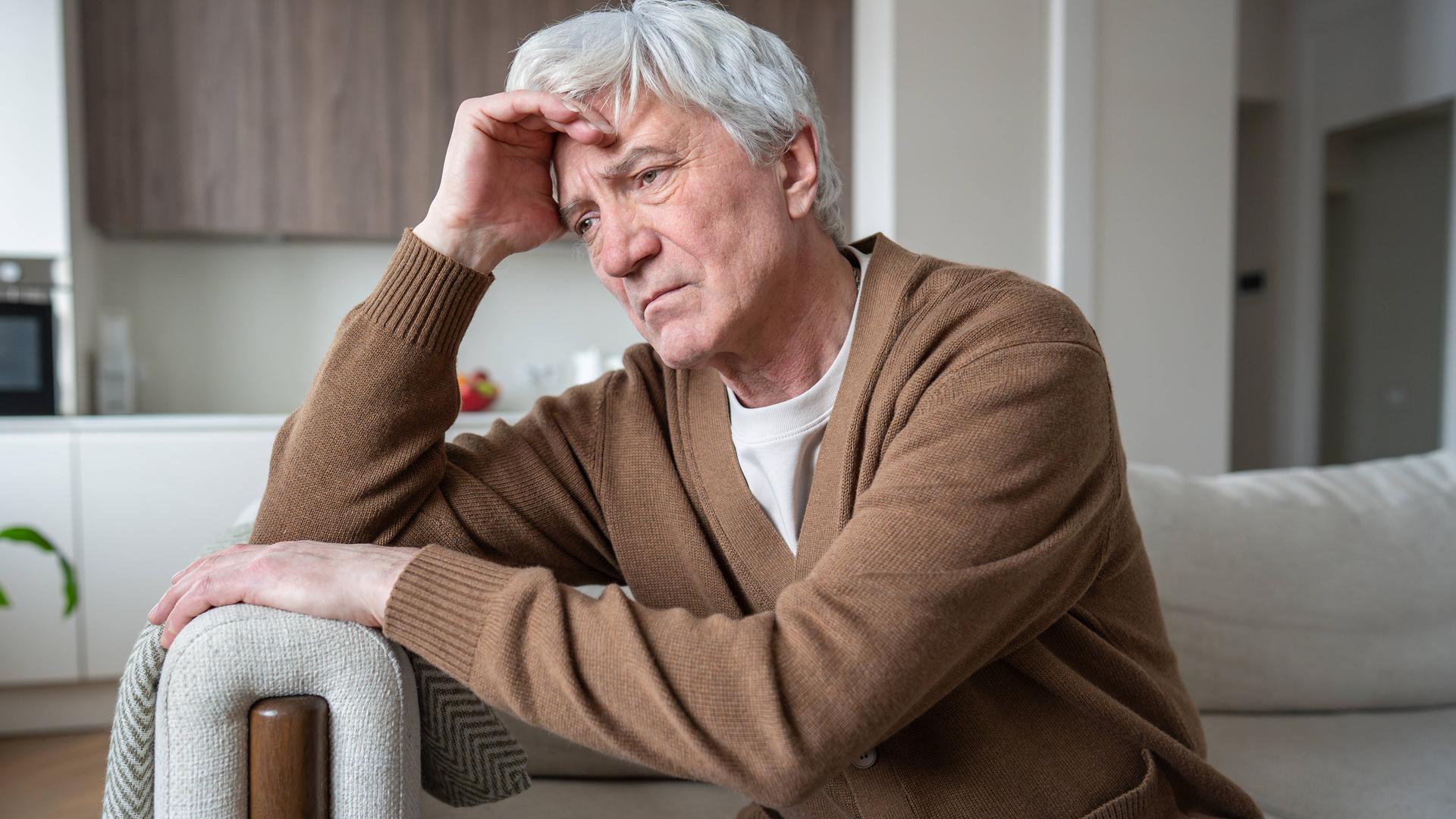 Älterer Mann sitzt auf dem Sofa, hält sich mit einer Hand den Kopf und blickt nachdenklich ins Leere.