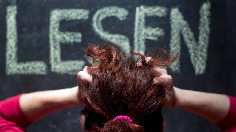 Eine Frau rauft sich vor einer Schultafel mit der Aufschrift "Lesen" die Haare.