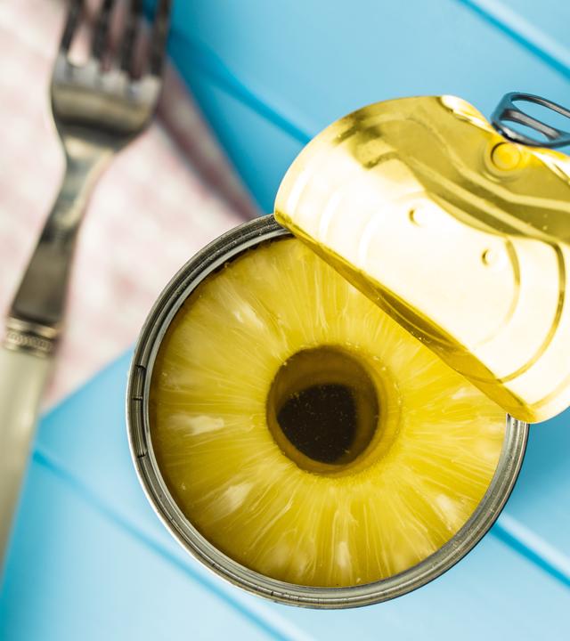 Auf einem blauen Tisch steht eine Dose in Scheiben geschnittene Ananas. Daneben liegt eine Gabel.