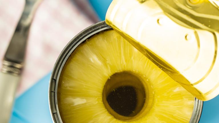 Auf einem blauen Tisch steht eine Dose in Scheiben geschnittene Ananas. Daneben liegt eine Gabel.