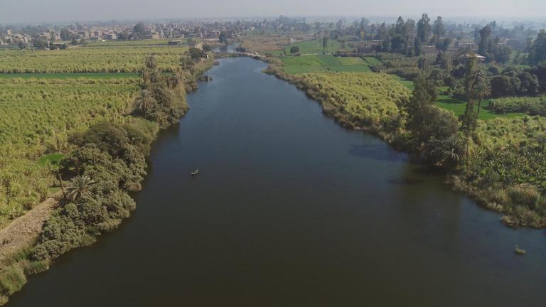 Ein breiter Fluss, auf dem ein Boot fährt, erstreckt sich zwischen dicht bewachsenen Ufern bis zum Horizont, wo eine Stadt an den Ufern liegt.