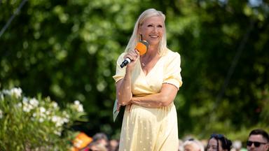 Andrea "Kiwi" Kiewel steht im Fernsehgarten, hält ein Mikrofon in der Hand und schaut lächelnd seitlich nach vorne.