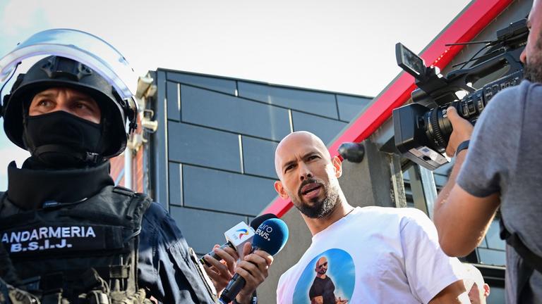 Andrew Tate (Mitte) spricht mit Journalisten, als er am 21. August 2024 von rumänischen Gendarmen zum Verhör vorgeführt wird, nachdem die Polizei ihr Haus in Bukarest zehn Stunden lang durchsucht hatte. 