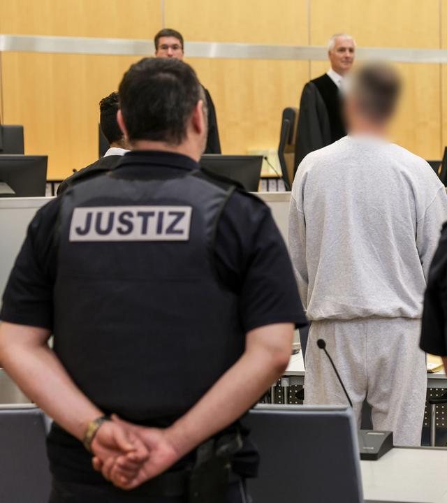Der Angeklagte (mittig) steht zwischen Justizangestellten im Gerichtssaal Düsseldorf am 04.05.2026 während des Prozesses um Politiker-Todeslisten, die er auf der Darknet-Plattform veröffentlichte.