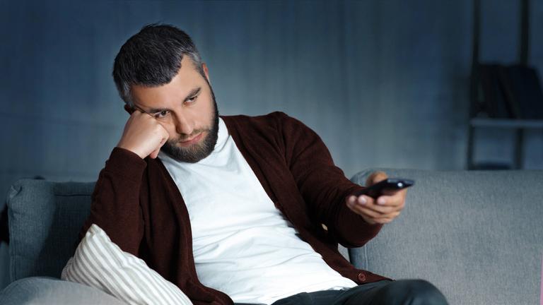 Mann sitzt auf dem Sofa und stützt seinen Kopf mit der Hand. In der anderen Hand hält er eine Fernbedienung.