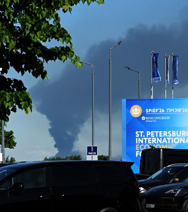 Am 03.06.2026 sind im russischen Sankt Petersburg Autos zu sehen, die in der Nähe von Bannern mit dem Logo des Internationalen Wirtschaftsforums von Sankt Petersburg (SPIEF) am Veranstaltungsort des Forums geparkt sind, während in der Ferne schwarzer Rauch aufsteigt. 