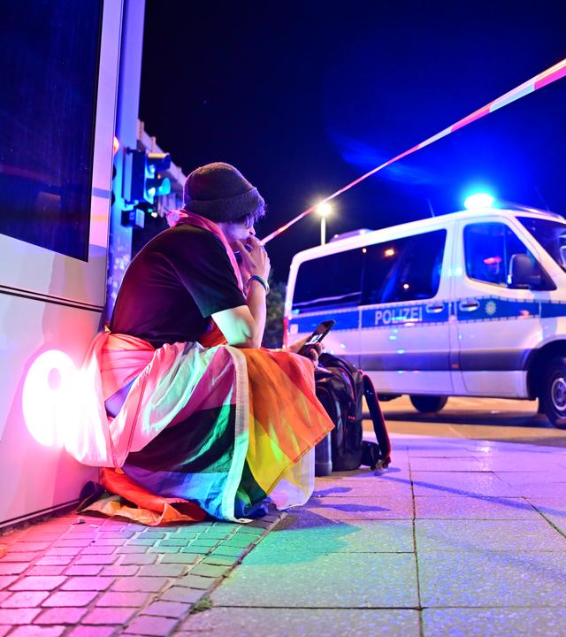Eine Person, in eine Pride-Fahne gehüllt, hockt neben einem Polizeifahrzeug nach dem Abbruch des Christopher Street Day (CSD) in Berlin.