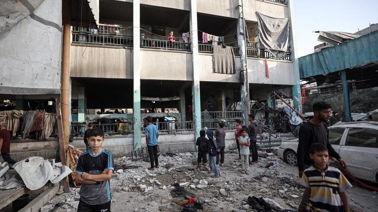 Ein zerstörtes Gebäude, was mal eine Schule war. Im Vordergrund stehen Kinder in den Trümmern.