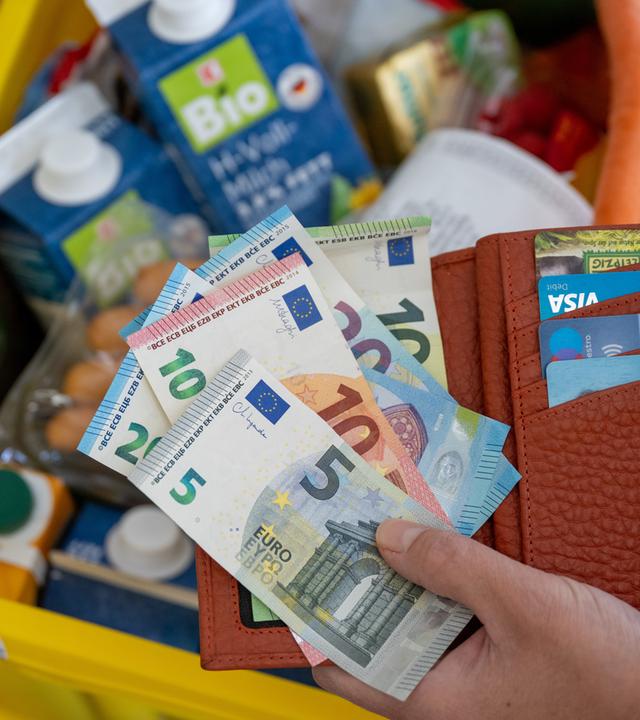 Eine Hand hält Geldscheine und einen Geldbeutel über einer Kiste Lebensmittel ins Bild.