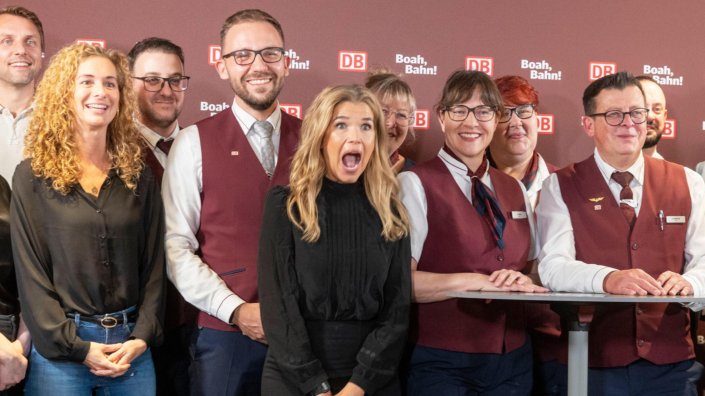 Anke Engelke in der Mitte des Bildes wie sie mit offenem Mund in die Kamera schaut, um sie herum mehrere Menschen mit DB-Klamotten.
