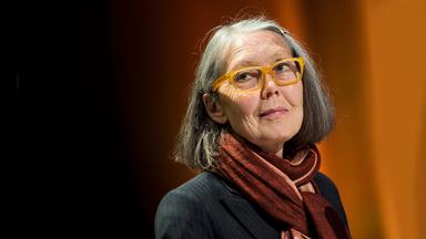 Porträtfoto einer älteren Frau mit schulterlangen grauen Haaren und einer Brille mit gelbem Kunststoffrahmen.