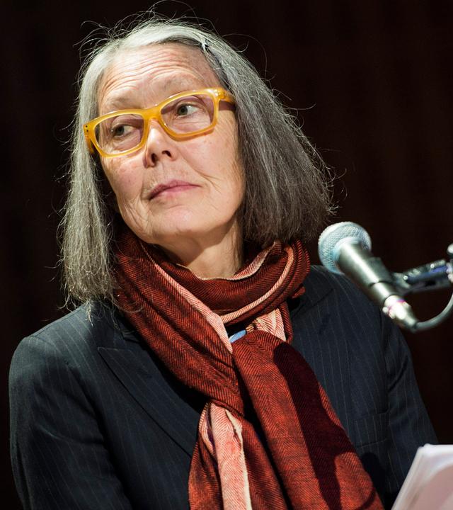 Porträtfoto einer älteren Frau mit schulterlangen grauen Haaren und einer Brille mit gelbem Kunststoffrahmen.
