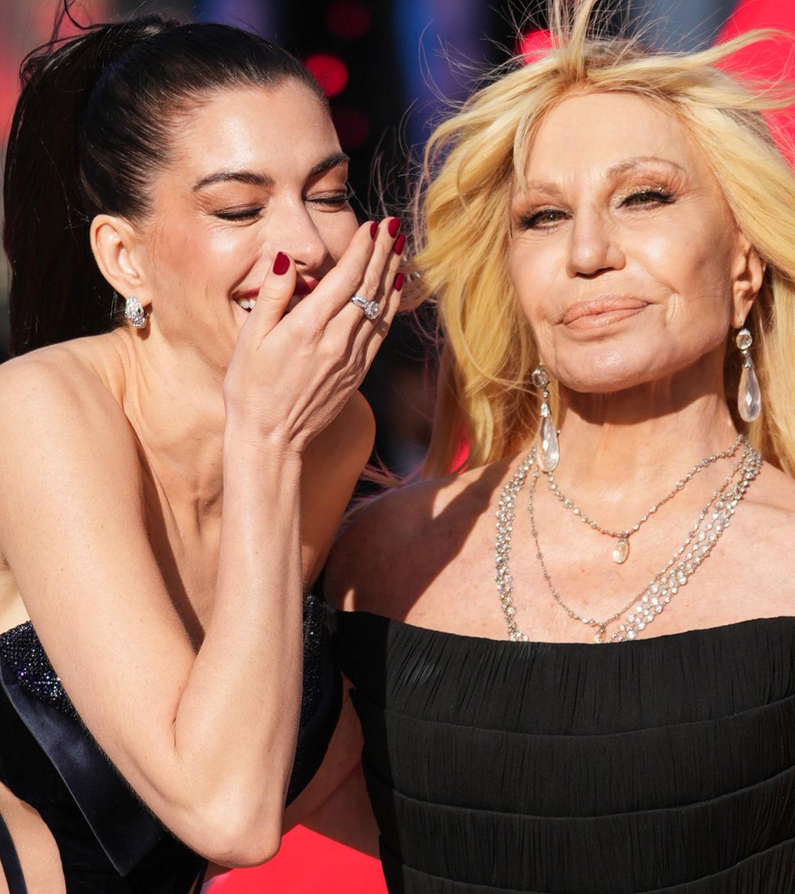 Anne Hathaway und Donatella Versace sind auf einer Filmpremiere zu sehen, Anne Hathaway kichert dabei und hält ihre Hand vor dem Mund.