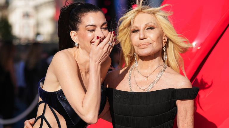 Anne Hathaway und Donatella Versace sind auf einer Filmpremiere zu sehen, Anne Hathaway kichert dabei und hält ihre Hand vor dem Mund.