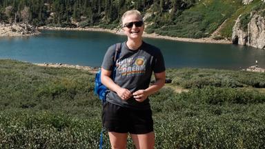 Anne McCarthy steht nach einer Wanderung vor einem See und lächelt in die Kamera.