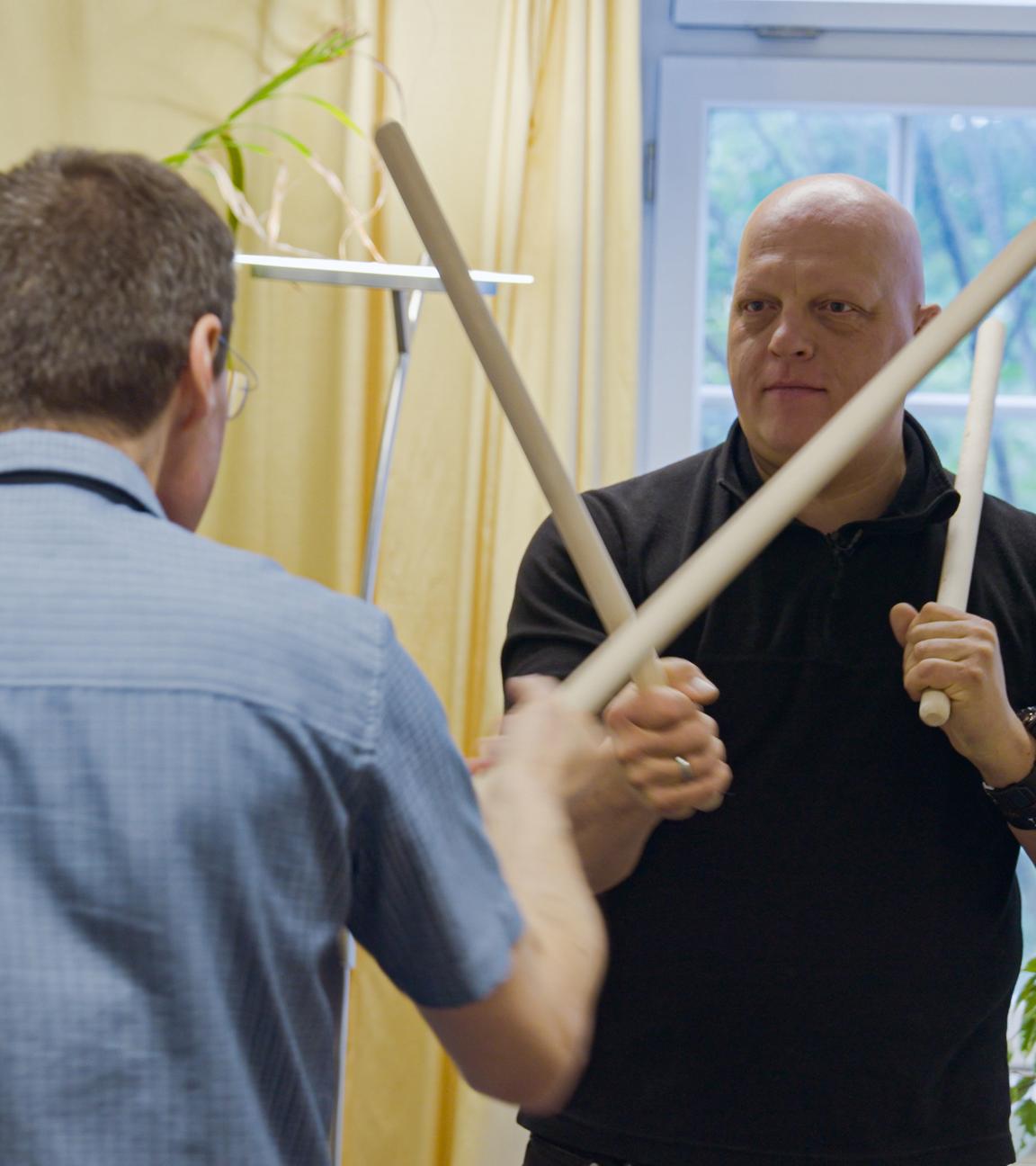 Zwei Männer stehen sich während einer Anti-Aggressionsübung mit jeweils zwei Stöcken gegenüber.