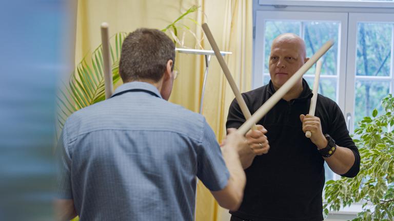 Zwei Männer stehen sich während einer Anti-Aggressionsübung mit jeweils zwei Stöcken gegenüber.
