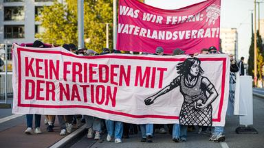 Menschen nehmen in Bremen an einer Demonstration der "Basisgruppe Antifaschismus Bremen" gegen den Tag der Deutschen Einheit teil. Auf Bannern sind die Schriftzüge "Was wir feiern: Vaterlandsverrat!" und "Kein Frieden mit der Nation" zu lesen. 