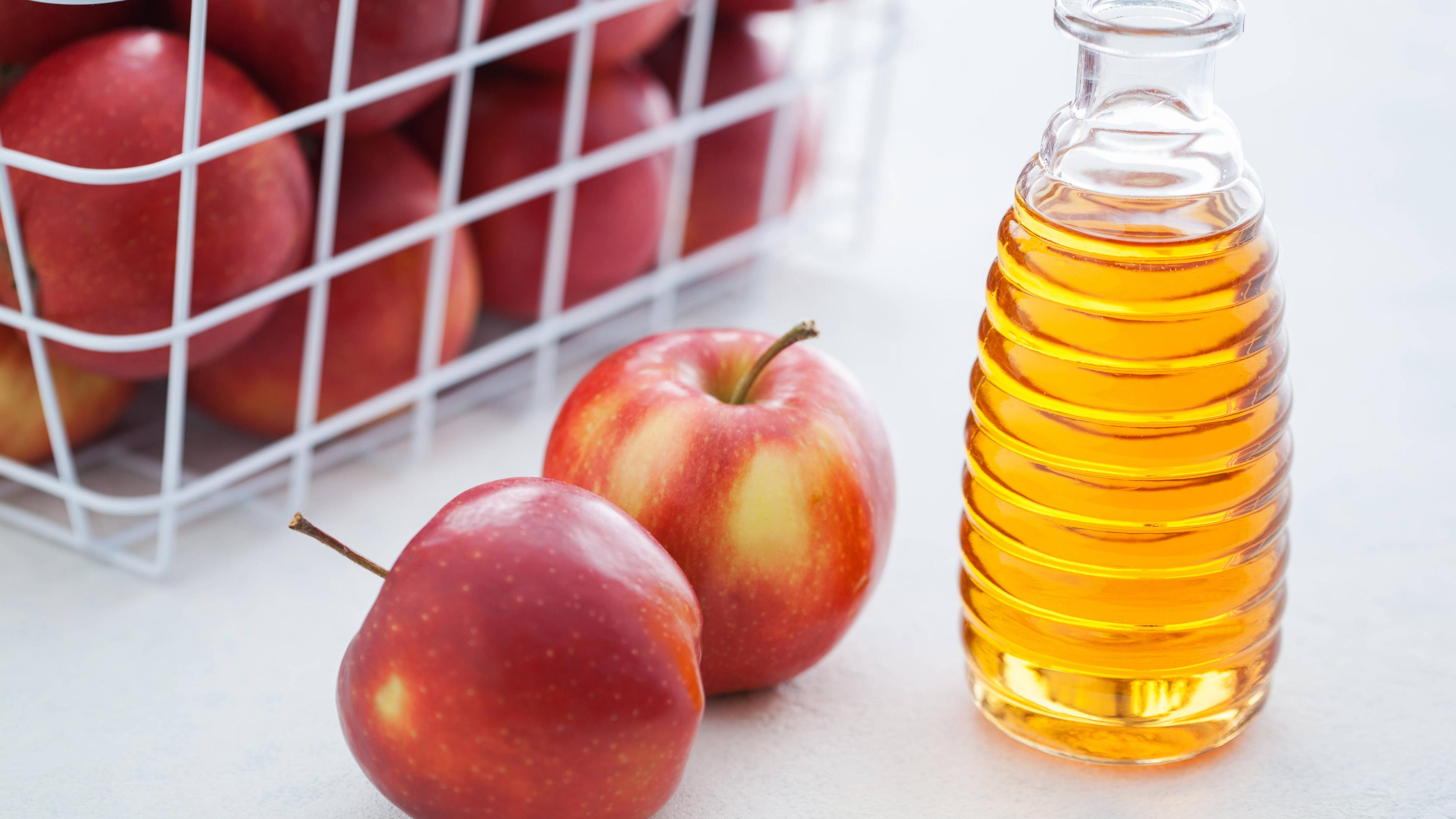 Eine Flasche Apfelessig steht neben Äpfeln mit einem Korb Äpfel im Hintergrund