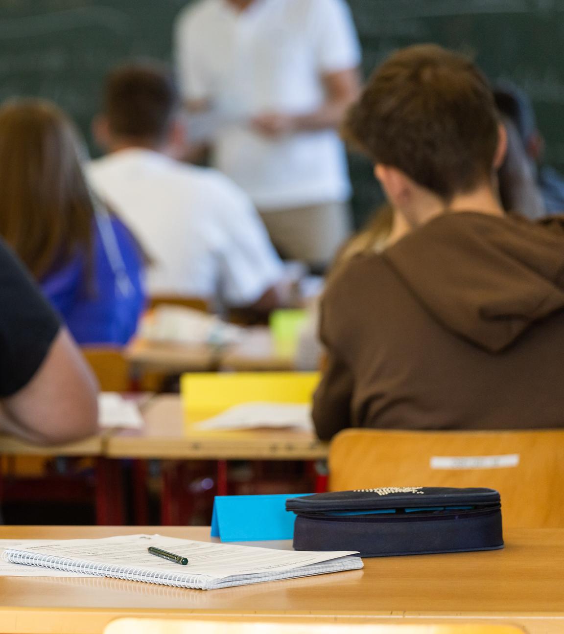 Baden-Württemberg, Heitersheim: Arbeitsmaterialien liegen auf einem Tisch in einem Klassenzimmer während dahinter Schüler und ihr Lehrer zu sehen sind.