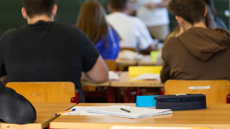 Baden-Württemberg, Heitersheim: Arbeitsmaterialien liegen auf einem Tisch in einem Klassenzimmer während dahinter Schüler und ihr Lehrer zu sehen sind.