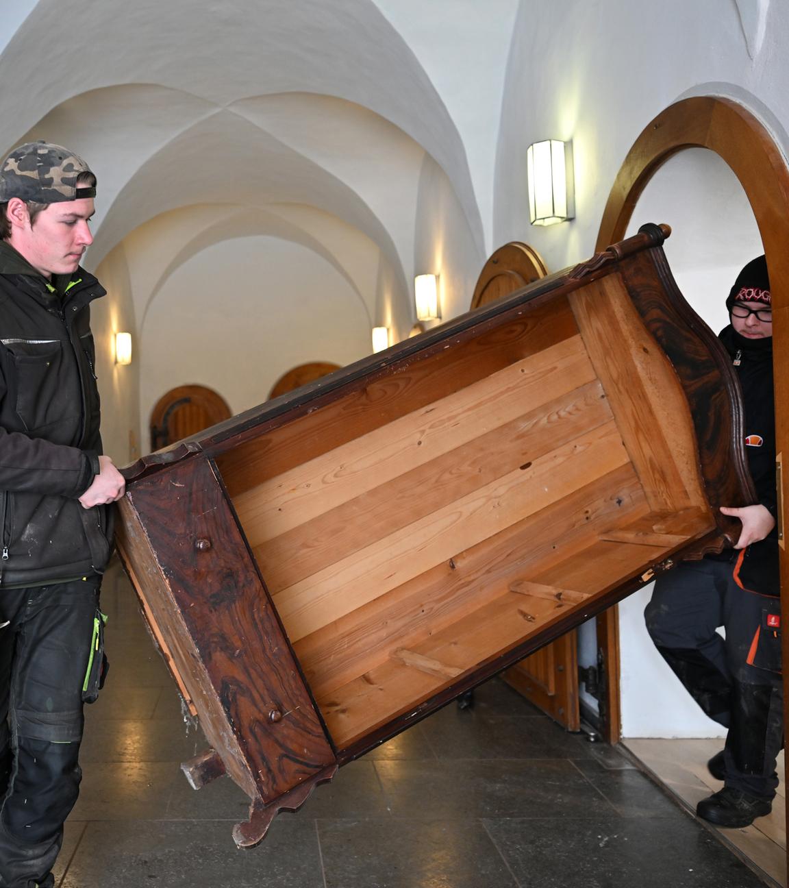Zwei junge Männer tragen ein aus Holz bestehendes Möbelstück.