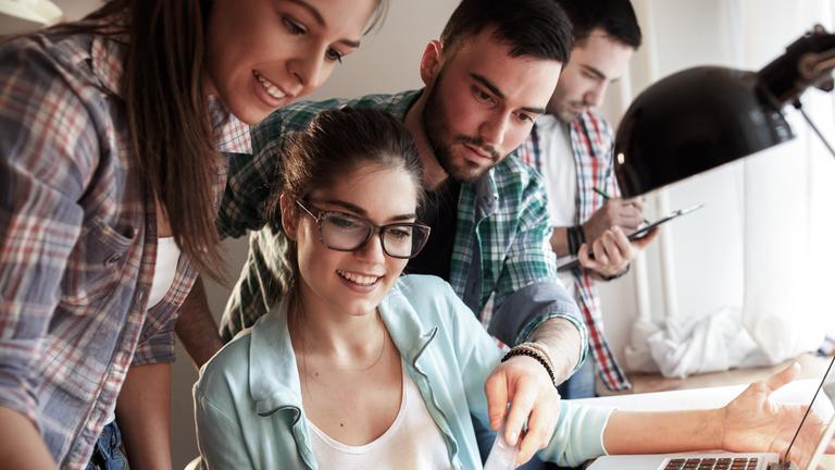 Vier junge Menschen beugen sich bei der Arbeit über einen Schreibtisch.