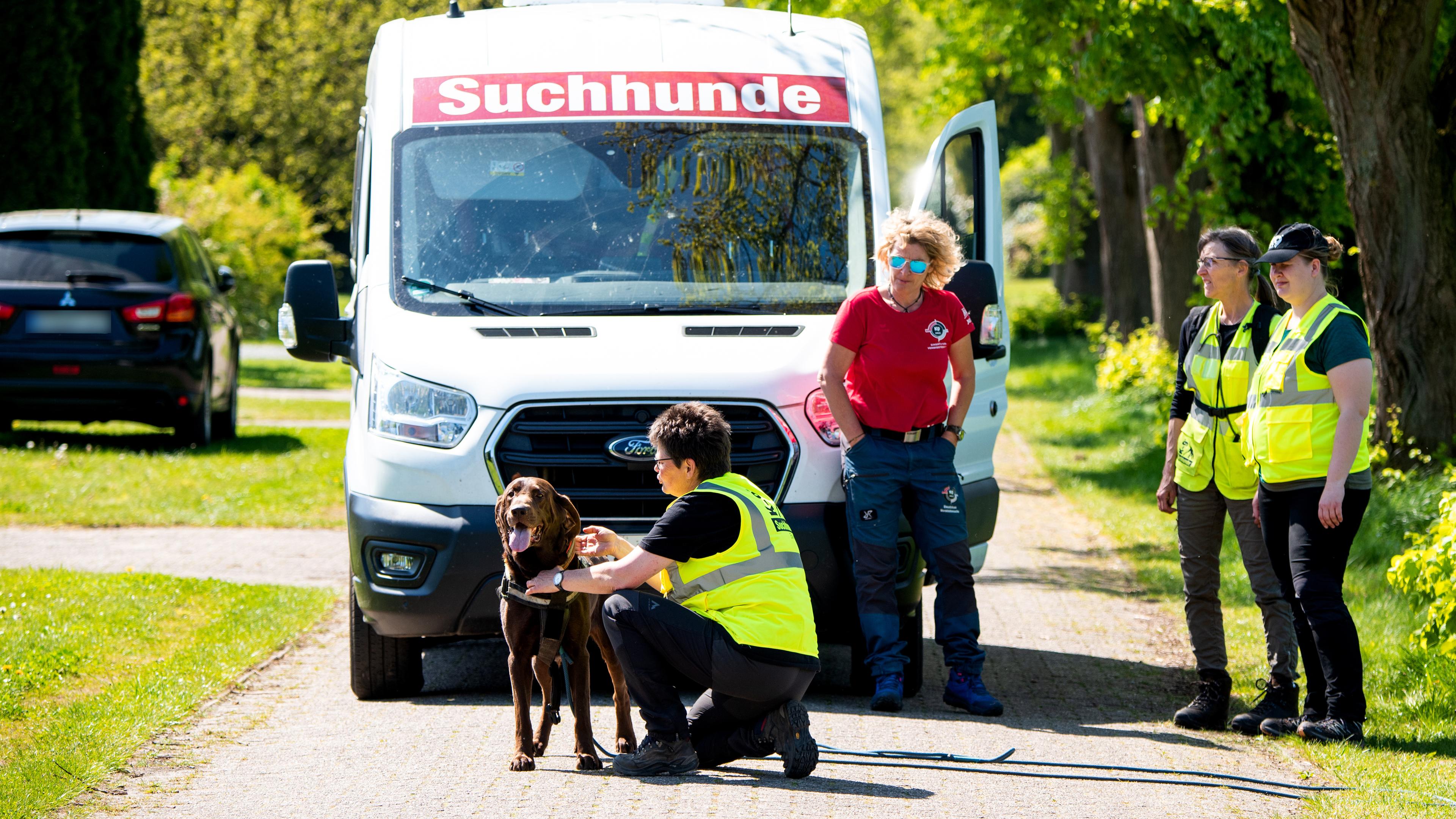 Ein Suchhund wird für die Suche nach einem vermissten Jungen in einem Wohngebiet vorbereitet. Mehrere Personen stehen um ihn, dahinter ein Auto.