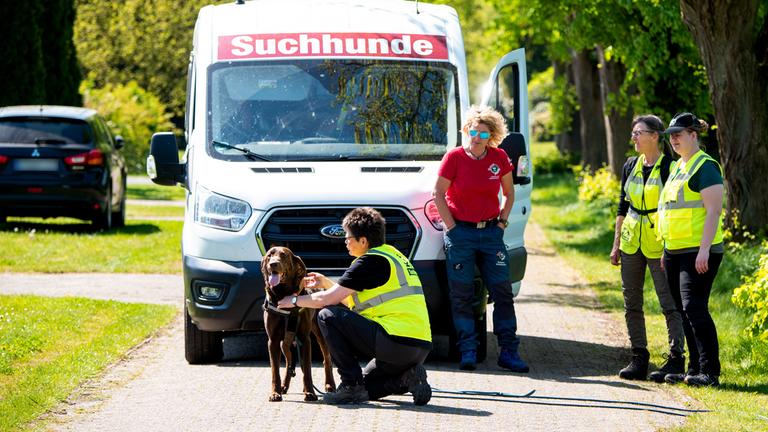 Ein Suchhund wird für die Suche nach einem vermissten Jungen in einem Wohngebiet vorbereitet. Mehrere Personen stehen um ihn, dahinter ein Auto.