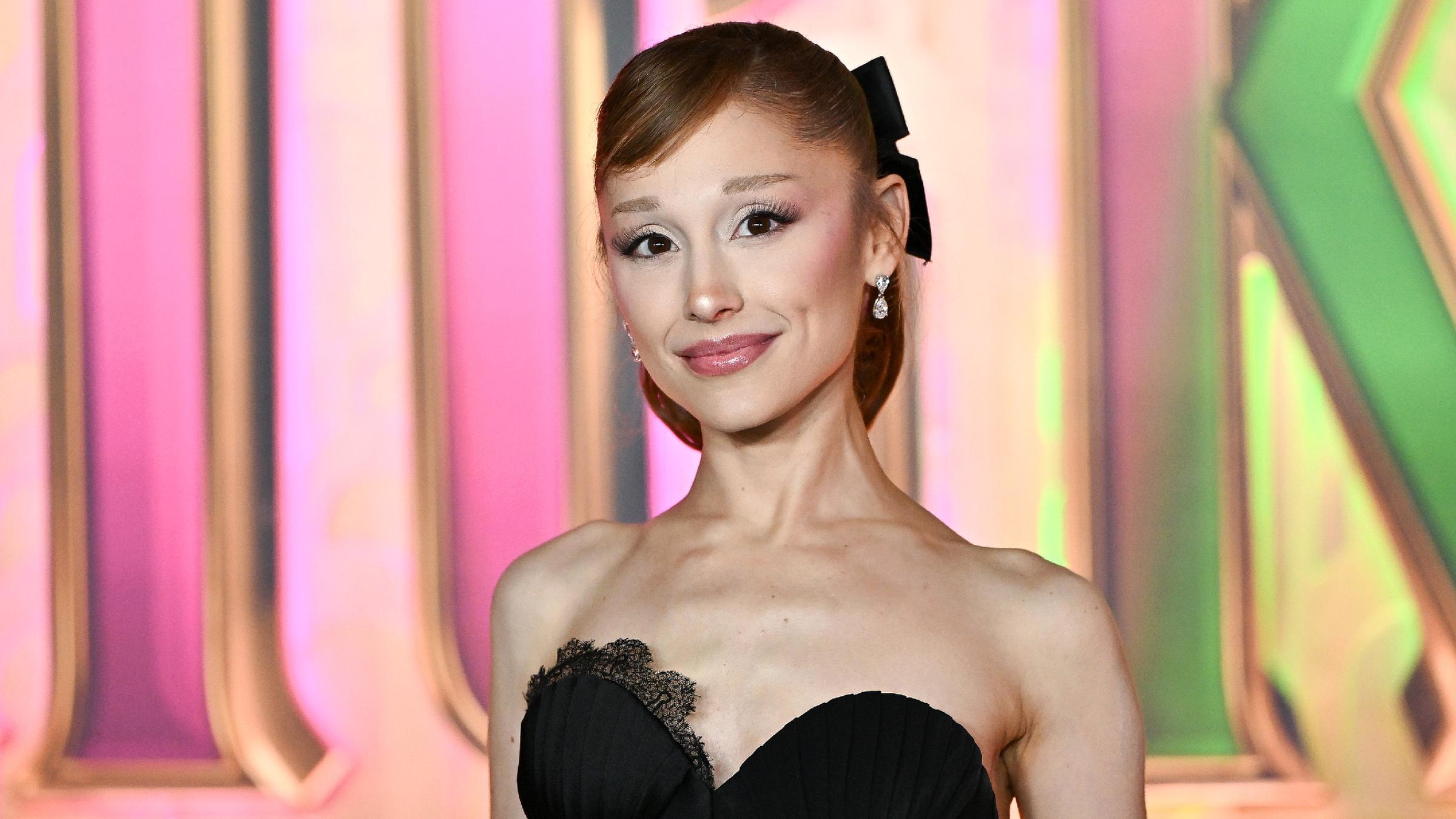 Die amerikanische Sängerin und Schausspielerin Ariana Grande steht in einem schwarzen Kleid vor einer bunten Wand