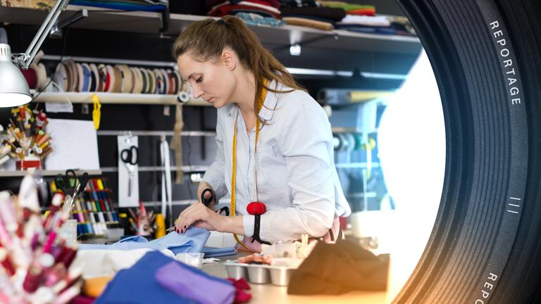 In einem Raum voller Stoffe, Schnüre und Fäden, zerschneidet eine Frau mit einem Maßband um den Hals konzentriert einen blauen Stoff.