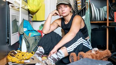 Luisa sitzt in einer Küche auf dem Boden und guckt Richtung Kamera. Um sie herum liegt ein Rucksack, ein Kuscheltier, ein Schulranzen.
