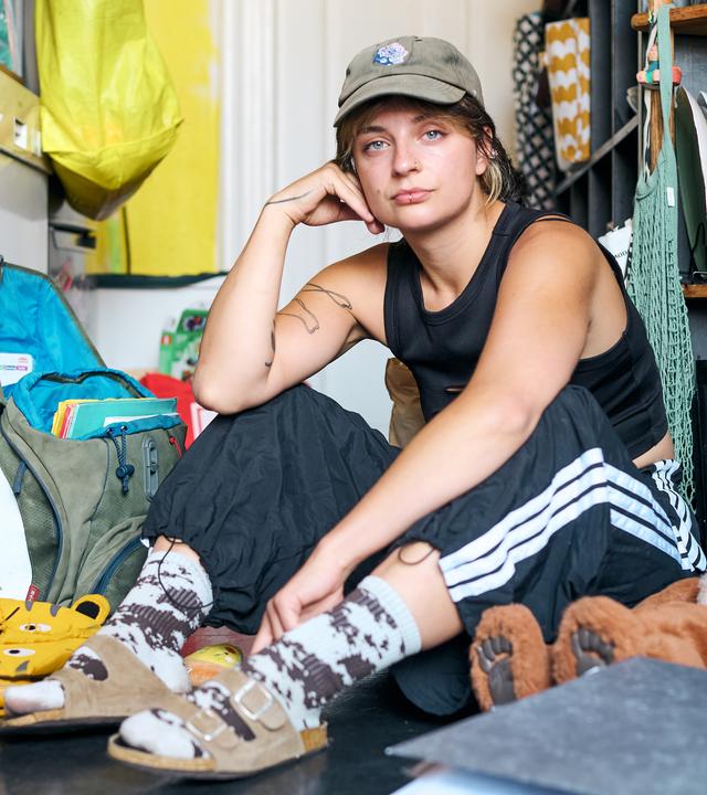 Luisa sitzt in einer Küche auf dem Boden und guckt Richtung Kamera. Um sie herum liegt ein Rucksack, ein Kuscheltier, ein Schulranzen.