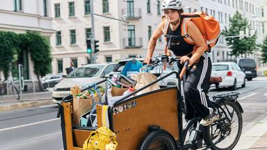 Luisa fährt mit einem Lastenrad auf einer Straße. Im Hintergrund sind Autos und Häuser zu sehen.